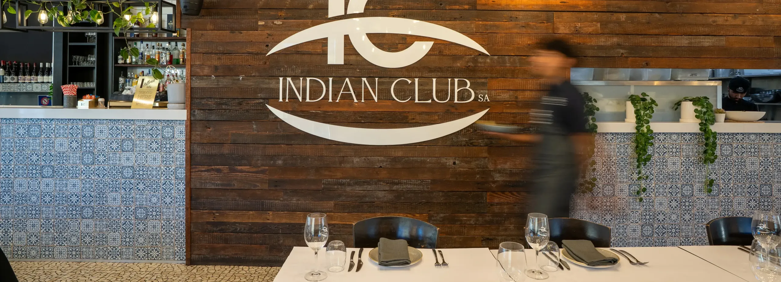 Waiter with food walking past an Indian Logo SA logo on the wall.